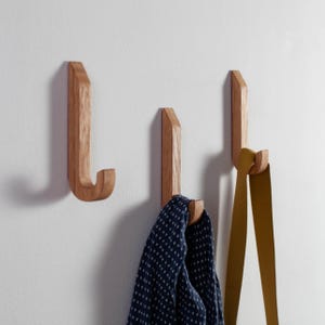 Könnte beinhalten: Drei Wandhaken aus Holz mit natürlicher Oberfläche sind an einer weißen Wand befestigt. Ein Haken hält einen marineblauen Schal mit weißen Punkten, ein anderer einen senffarbenen Lederriemen. Die Haken haben ein schlichtes, modernes Design.