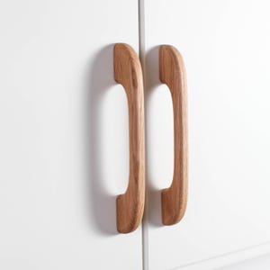 Könnte beinhalten: Zwei Holzschrankgriffe mit geschwungenem Design. Die Griffe bestehen aus hellfarbigem Holz und sind an einer weißen Schranktür befestigt. Die Griffe sind vertikal angeordnet und haben eine glatte, polierte Oberfläche.