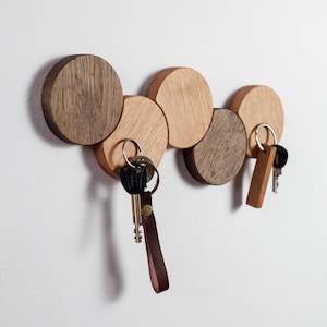 Könnte beinhalten: Ein hölzerner Schlüsselhalter mit sechs kreisförmigen Holzstücken in verschiedenen Brauntönen. Schlüssel mit Metallringen und Leder- oder Holzanhängern hängen an dem Schlüsselhalter. Der Schlüsselhalter ist an einer weißen Wand befestigt.