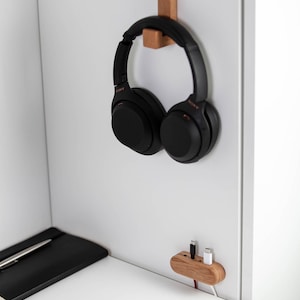 May include: Black over-ear headphones hanging on a wooden hook mounted on a white wall. A wooden charging station with two cables is also mounted on the wall. A black notebook and pen are on a white desk.