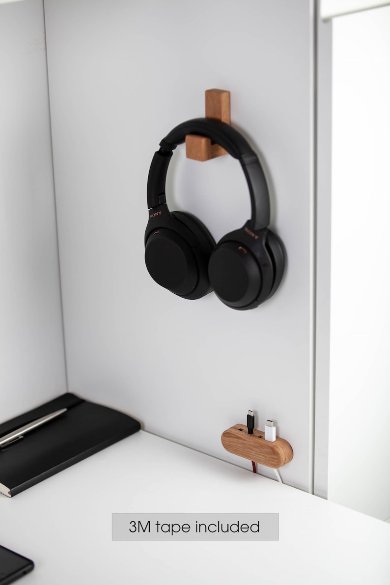 May include: Black over-ear headphones hanging on a wooden hook mounted on a white wall. A wooden charging station with two cables is also mounted on the wall. A black notebook and pen are on a white desk.