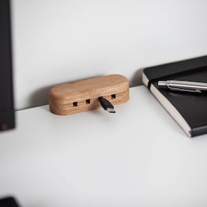 May include: A wooden USB hub with a light brown finish. The hub has four square ports and a black USB-C cable plugged into one. A black notebook and a silver pen are in the background.