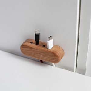 May include: A wooden cable organizer with a curved design. It features three holes for holding cables, with a black and a white cable inserted. The organizer is made of light brown wood and is mounted on a white surface.