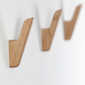 Op de afbeelding: Drie houten wandhaken met een natuurlijke houtnerf afwerking zijn op een witte muur gemonteerd. De haken hebben een modern, minimalistisch ontwerp, met een gebogen vorm en een platte basis voor montage. De haken zijn uit elkaar geplaatst.