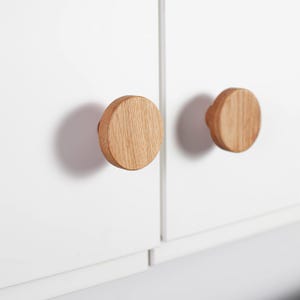 Puede incluir: Dos pomos redondos de madera clara están montados en una puerta de armario blanca. Los pomos tienen un patrón de vetas de madera natural y están espaciados uniformemente. La puerta del armario tiene un diseño limpio y moderno.