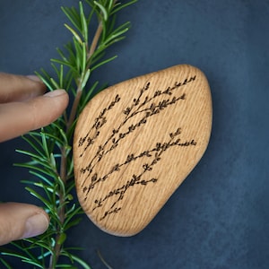 May include: A light-coloured wooden decorative stone with a floral design. The stone is a natural shape and has a smooth surface. The floral design is etched into the wood in a dark brown colour. A sprig of rosemary is next to the stone.