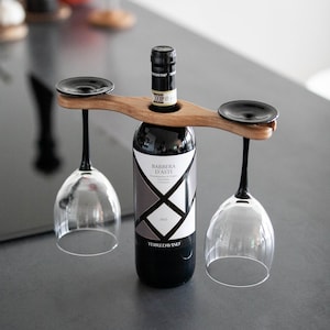 Könnte beinhalten: Ein Weinhalter aus Holz mit zwei Weingläsern mit schwarzem Stiel. Der Halter hat auch zwei schwarze runde Teller. Eine Flasche Rotwein befindet sich in der Mitte des Halters. Die Weinflasche hat ein schwarz-weißes Etikett.