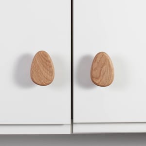 Könnte beinhalten: Zwei eiförmige Holzschrankknöpfe. Die Knöpfe sind aus hellem Holz gefertigt und an einer weißen Schranktür befestigt. Die Holzmaserung ist sichtbar und verleiht den Knöpfen Textur. Schlichtes, modernes Design. Ca. 5 cm lang.