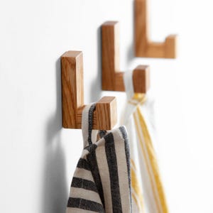 Könnte beinhalten: Drei Wandhaken aus Holz mit natürlicher Oberfläche sind an einer weißen Wand befestigt. Zwei Haken halten gestreifte Handtücher, eines schwarz-weiß, das andere gelb-weiß. Die Haken sind aus massivem Holz gefertigt und haben ein schlichtes, modernes Design.