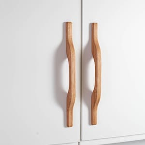 Könnte beinhalten: Zwei Holzschrankgriffe mit geschwungenem Design an einer weißen Schranktür. Die Griffe sind aus hellem Holz gefertigt und haben eine glatte, organische Form, die einen Hauch von natürlicher Eleganz verleiht.