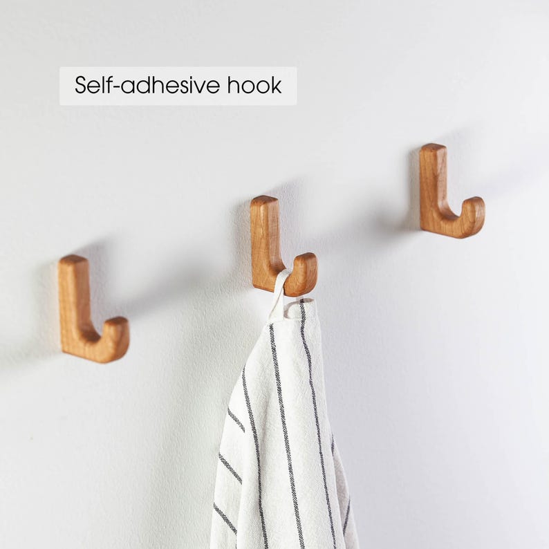 May include: Three wooden self-adhesive hooks mounted on a white wall. One hook has a white and black striped kitchen towel hanging from it.