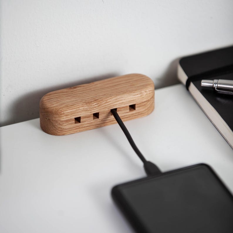 May include: A wooden charging station with three ports and a black charging cable. The station is made of light-colored wood and is connected to a black smartphone. The design is minimalist and modern.