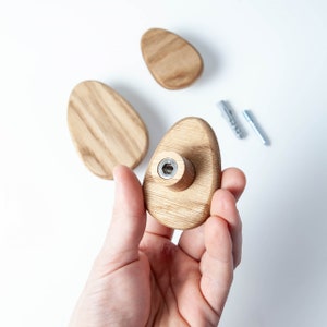 Pu&ograve; includere: Un gancio da parete in legno a forma ovale con una vite in metallo al centro. Il gancio &egrave; tenuto in una mano.