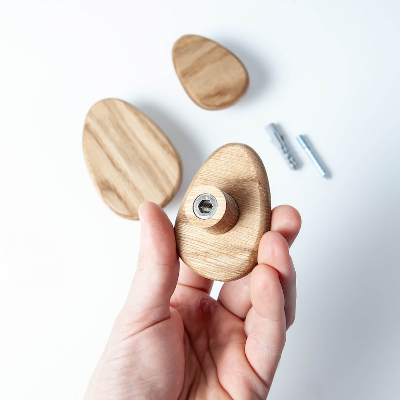 Pu&ograve; includere: Un gancio da parete in legno a forma ovale con una vite in metallo al centro. Il gancio &egrave; tenuto in una mano.