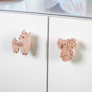 Könnte beinhalten: Zwei Holzknaufgriffe, einer in Form eines Rehs und der andere in Form eines Eichhörnchens. Beide sind in einem hellen Holztönen lackiert.