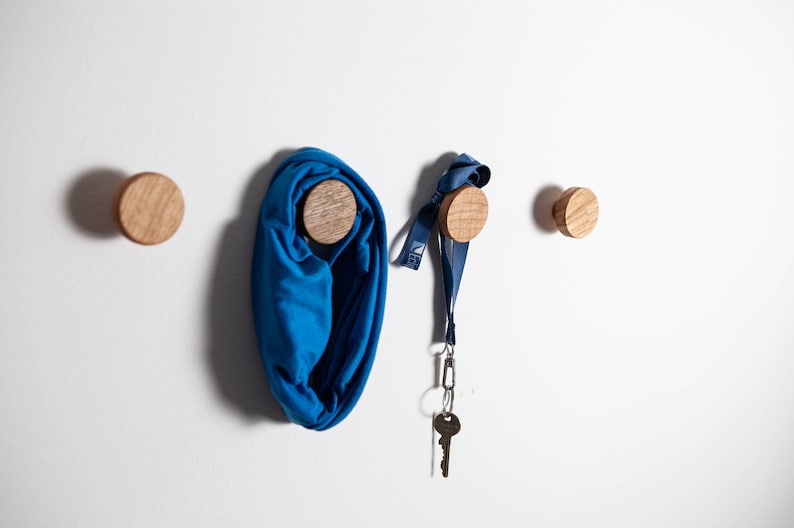 May include: Four wooden knobs mounted on a white wall. A blue scarf is hanging from one knob, and a blue lanyard with a key is hanging from another knob.