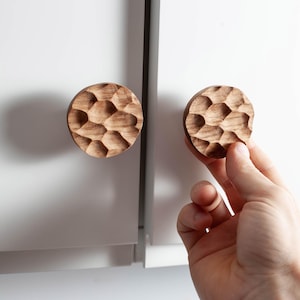 Peut inclure: Deux boutons de meuble ronds en bois avec une surface texturée et sculptée. L'un est fixé à une porte d'armoire blanche, l'autre est tenu par une main. Les boutons sont d'une couleur bois naturelle et chaleureuse, ajoutant une touche rustique à l'armoire. Environ 5 cm de diamètre.