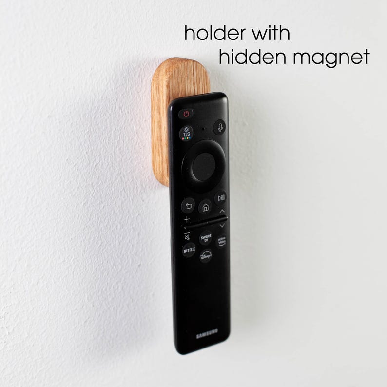 May include: A wooden remote control holder with a hidden magnet. The holder is attached to a white wall and a black remote control is attached to the holder.