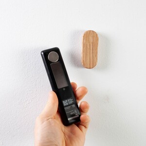May include: A black handheld electronic device with a small screen and a silver button. The device is being held in a person's hand. A small wooden oval hook is mounted on a white wall.