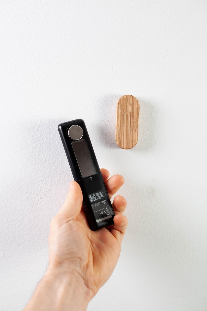 May include: A black handheld electronic device with a small screen and a silver button. The device is being held in a person's hand. A small wooden oval hook is mounted on a white wall.