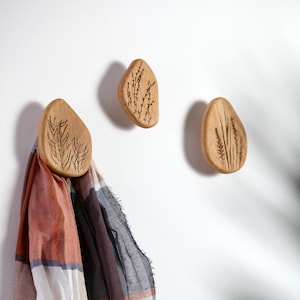 Könnte beinhalten: Drei Wandhaken aus Holz mit natürlicher Oberfläche und eingravierten botanischen Designs. Ein Haken hält einen Schal mit rot-orange-blauem Karomuster. Die Haken sind an einer weißen Wand befestigt und bieten eine dekorative und funktionale Aufbewahrungslösung.