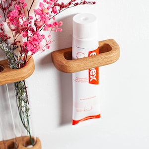 Op de afbeelding: Een houten tandenborstelhouder met een tube tandpasta en een vaas met roze bloemen. De tandpastatube is wit met oranje accenten en tekst. De houder is aan een witte muur bevestigd.