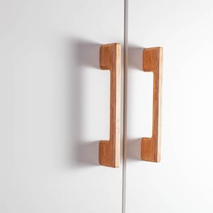 Könnte beinhalten: Zwei Holzschrankgriffe mit hellbraunem Finish sind auf einer weißen Oberfläche montiert. Die Griffe haben eine rechteckige Form mit einem Griffausschnitt und sind für einen einfachen Griff konzipiert.