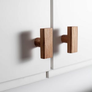 Könnte beinhalten: Zwei Holzschrankgriffe mit rechteckigem Design. Die Griffe sind aus hellbraunem Holz gefertigt und an einer weißen Schranktür montiert. Die Griffe haben ein T-förmiges Design.
