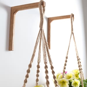 Könnte beinhalten: Zwei Wandhalterungen aus Holz für Pflanzen mit Makramee-Seil-Pflanzenhaltern. Die Pflanzenhalter sind mit bunten Blumen gefüllt, darunter gelbe und violette Blüten. Die Holzhalterungen sind hellbraun.