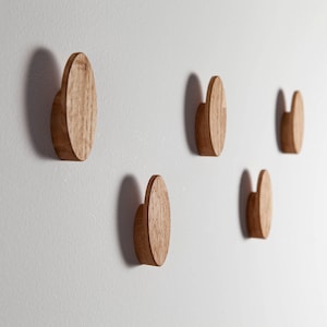 Könnte beinhalten: Fünf runde, hölzerne Wandhaken, die an einer weißen Wand befestigt sind. Die Haken sind alle gleich groß und haben die gleiche Form und sind versetzt angeordnet.