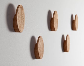 Op de afbeelding: Vijf houten wandhaken in de vorm van een afgeronde rechthoek, gemonteerd op een witte muur. De haken zijn allemaal even groot en hebben dezelfde vorm, en ze zijn in een verspringend patroon gerangschikt.