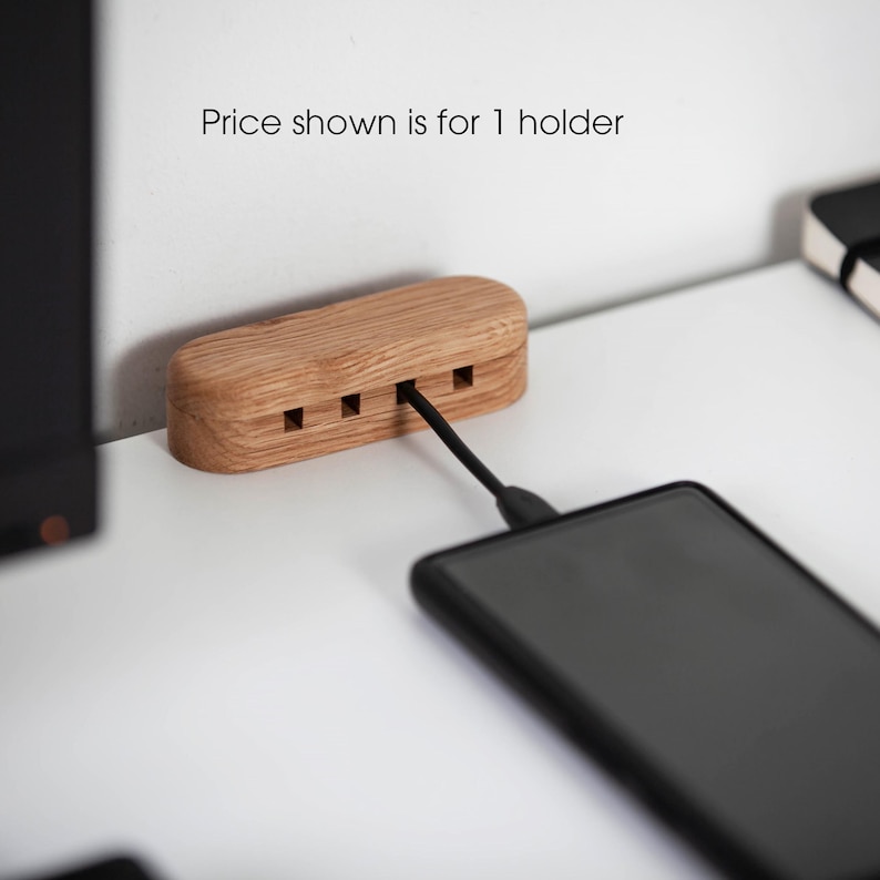 May include: A wooden phone charging station with three slots, connected to a black smartphone via a black charging cable. The station is made of light-colored wood and is rectangular with rounded edges. The text "Price shown is for 1 holder" is visible.