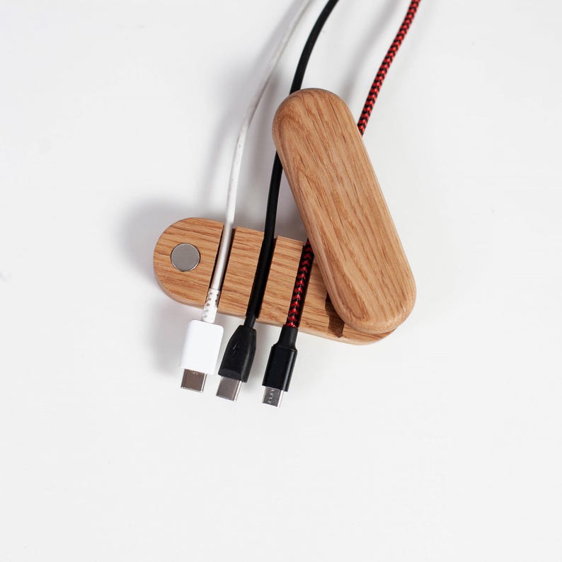 May include: A wooden cable organizer with three charging cables: white, black, and red. The organizer is made of light-colored wood and has a magnetic closure. The cables are plugged into various devices.