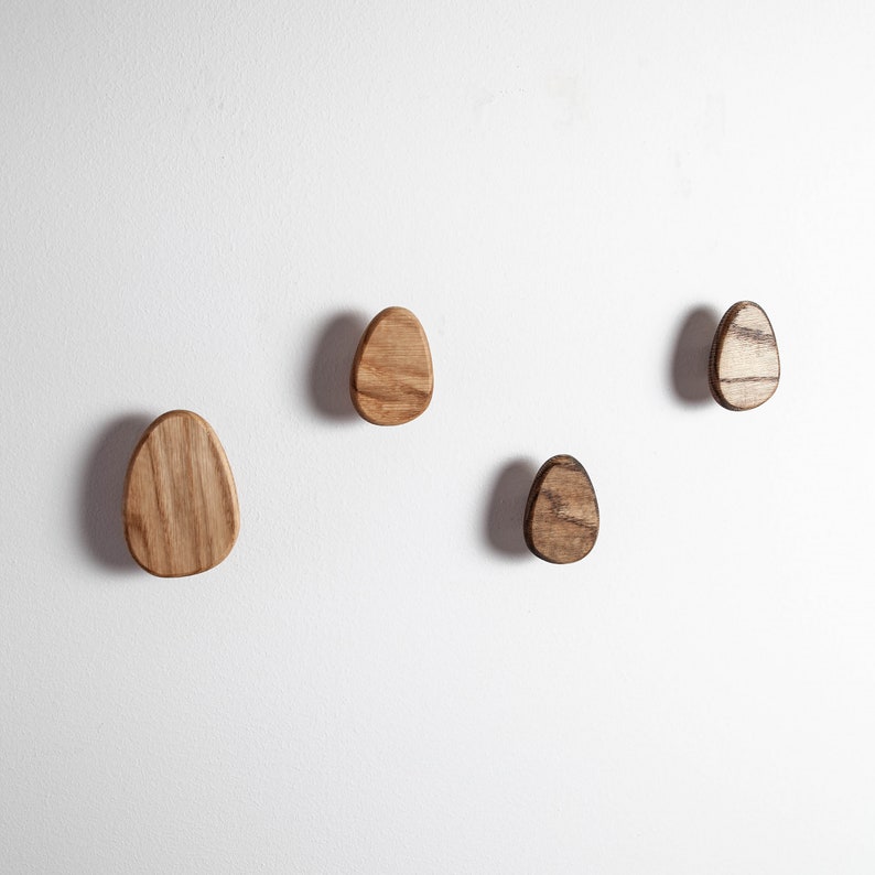 Pu&ograve; includere: Quattro ganci da parete in legno a forma di uovo. I ganci sono di varie tonalit&agrave; di marrone e sono montati su una parete bianca.