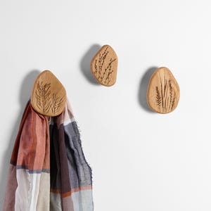 Op de afbeelding: Drie houten wandhaken met een natuurlijke afwerking en gegraveerde botanische ontwerpen. Eén haak houdt een kleurrijke sjaal vast. De haken zijn kiezelvormig en voorzien van verschillende plantenillustraties, die een vleugje natuur toevoegen.