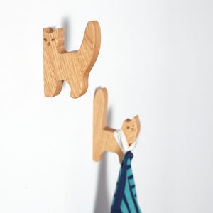 Op de afbeelding: Twee houten katvormige wandhaken. De haken zijn gemaakt van lichtgekleurd hout en hebben een eenvoudig kattenontwerp met een staart en kleine gezichtsdetails. Eén haak houdt een blauw-wit gestreepte handdoek vast.