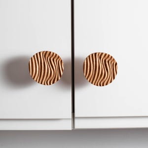 Op de afbeelding: Twee ronde houten kastknoppen met een uitgesneden, golvend patroon. De knoppen zijn lichtbruin van kleur en zijn gemonteerd op een witte kastdeur. De knoppen hebben een diameter van ongeveer 5 cm.