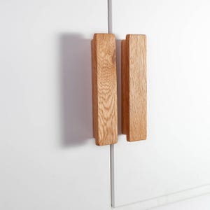 Könnte beinhalten: Zwei rechteckige Holzschrankgriffe mit natürlicher Holzmaserung sind an einer weißen Schranktür befestigt. Die Griffe sind vertikal ausgerichtet und haben ein schlichtes, modernes Design. Die Schranktür ist geschlossen.