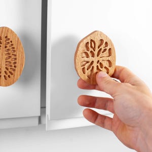 Könnte beinhalten: Schrankknöpfe aus Holz mit geschnitztem Design. Ein Knopf wird von einer Hand gehalten, während ein anderer an einer weißen Schranktür befestigt ist. Die Knöpfe haben eine natürliche Holzoberfläche und ein einzigartiges, dekoratives Muster.