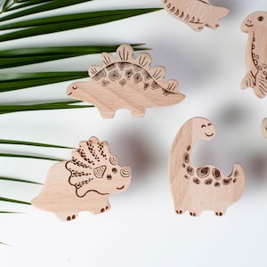 Puede incluir: Juguetes de dinosaurios de madera en varias formas, incluyendo un Estegosaurio y un Brontosaurio, dispersos sobre una superficie blanca. Cada juguete tiene detalles grabados y un acabado de madera natural. Hojas verdes en el fondo.
