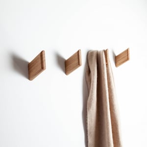 Peut inclure: Trois patères murales en bois en forme de losange sont fixées sur un mur blanc. Une écharpe beige est suspendue à l'un des crochets. Les crochets sont en bois clair.