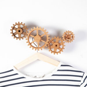 Könnte beinhalten: Haken aus Holz in Form von Zahnrädern, die an einer weißen Wand montiert sind. Ein gestreiftes Hemd hängt an einem Holzbügel an den Haken.