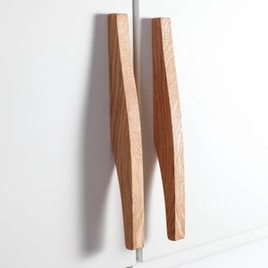Könnte beinhalten: Zwei Holzschrankgriffe mit hellbraunem Finish. Die Griffe haben ein geometrisches, eckiges Design und sind an einer weißen Schranktür montiert. Die Griffe sind aus Holz und haben eine natürliche Maserung.