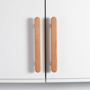 Könnte beinhalten: Zwei Holzschrankgriffe mit hellbraunem Finish sind an einem weißen Schrank montiert. Die Griffe sind vertikal, rechteckig und haben abgerundete Enden. Die Schranktüren sind geschlossen, mit einer dünnen schwarzen Linie dazwischen.