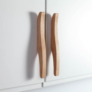 Könnte beinhalten: Zwei Holzschrankgriffe mit hellbraunem Finish sind an einer weißen Schranktür montiert. Die Griffe haben ein gebogenes, ergonomisches Design und sind aus massivem Holz gefertigt. Die Schranktüren sind geschlossen und die Griffe vertikal angeordnet.