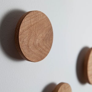 May include: Three round wooden wall hooks mounted on a white wall. The hooks are made of natural wood with a light brown color and a slightly rough texture.