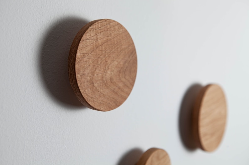 May include: Three round wooden wall hooks mounted on a white wall. The hooks are made of natural wood with a light brown color and a slightly rough texture.