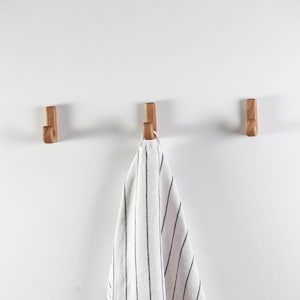 May include: Three wooden hooks mounted on a white wall with a striped kitchen towel hanging from the center hook.