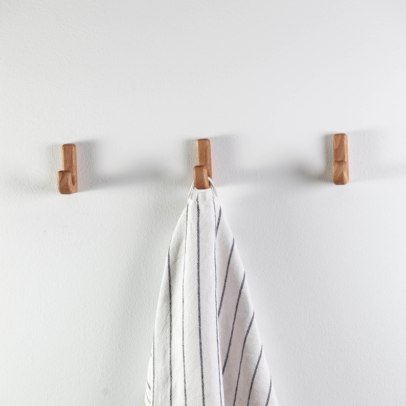May include: Three wooden hooks mounted on a white wall with a striped kitchen towel hanging from the center hook.