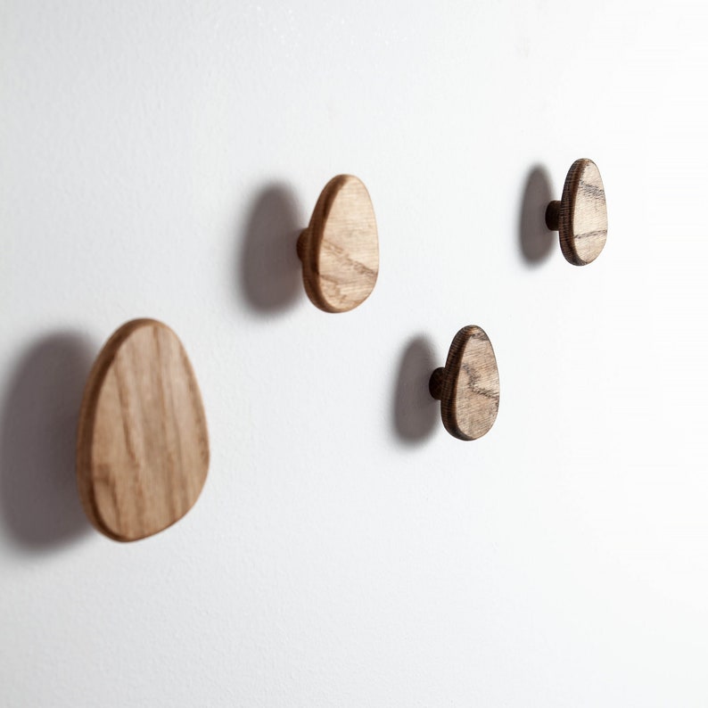 Pu&ograve; includere: Quattro ganci da parete in legno a forma ovale, montati su una parete bianca. I ganci sono in legno naturale di colore marrone chiaro con una venatura marrone leggermente pi&ugrave; scura.